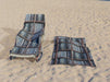 a towel with image of vinyl records stacked along shelves, the towel on a lounger on a beach with an identical towel on the sand beside it