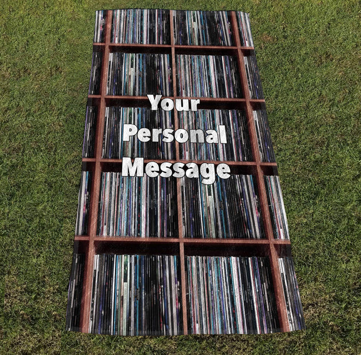 a towel with image of vinyl records stacked along shelves with a personal printed message, the towel laid out on grass