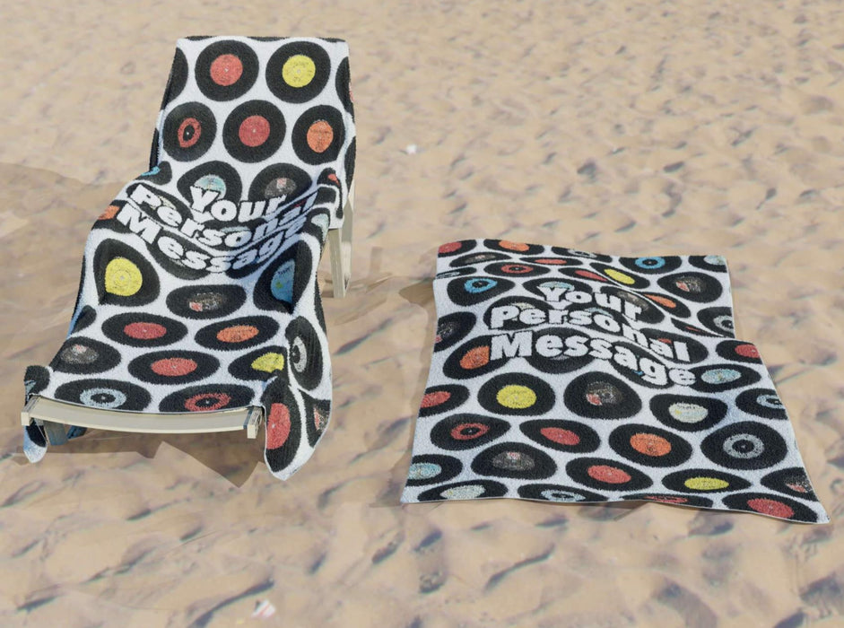 a towel with image of 7 inch vinyl records in a mosaic pattern along with a personal message printed, the towel on a lounger on a beach with an identical towel on the sand beside it